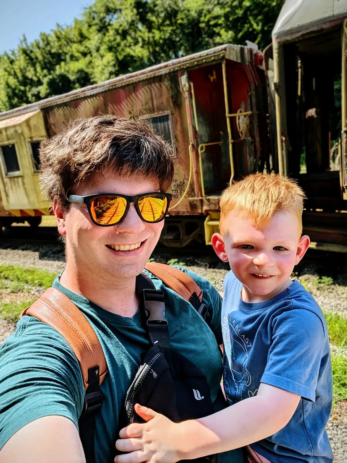 Nathan carrying a small child on a sunny day in front of old railroad cars.