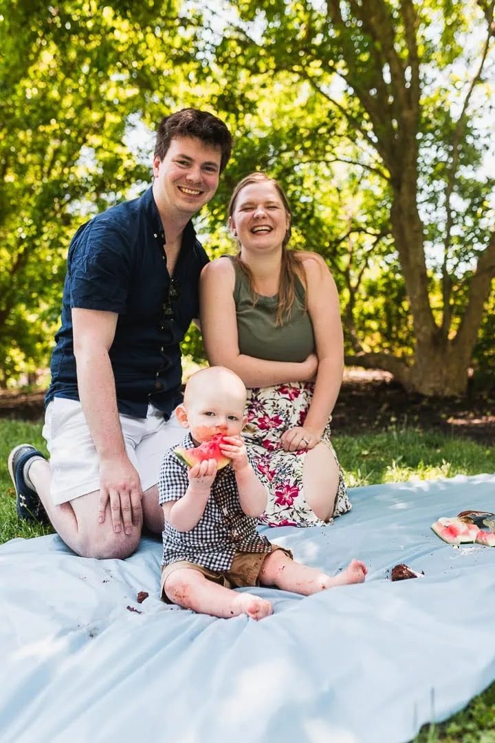 Nathan with his family on a picnic blanket in a park, a young child eating watermelon.