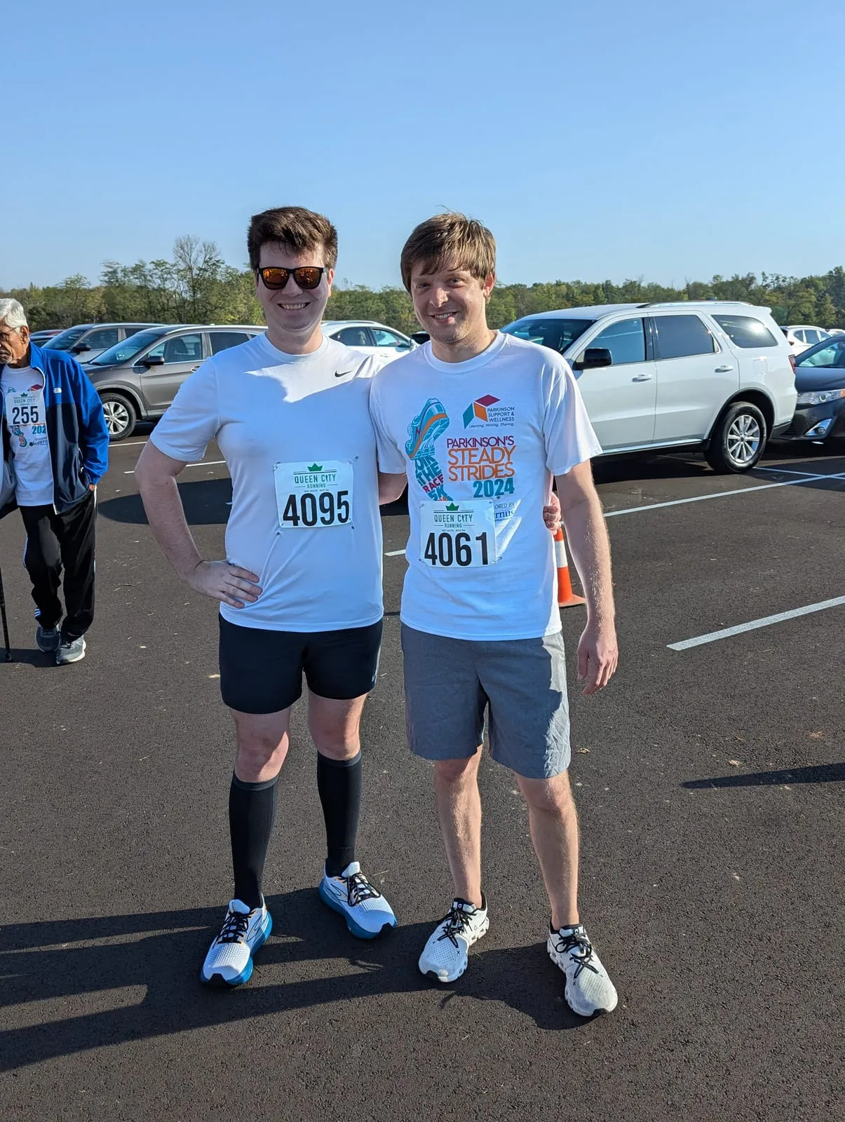 Nathan and a friend at the end of a charity 5K, race bibs on, in a sunny parking lot.