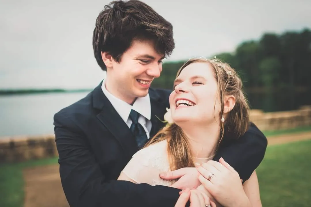 Nathan and his wife laughing together on their wedding day, beside a lake.
